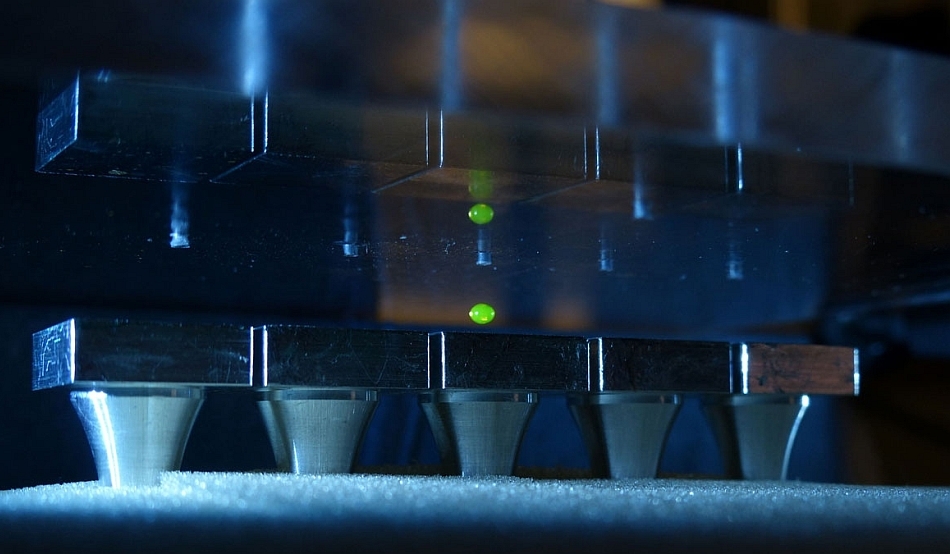 O picătură de lichid levitează între platformele ce emit sunete (jos) şi suprafaţa de sus ce reflectă sunetele